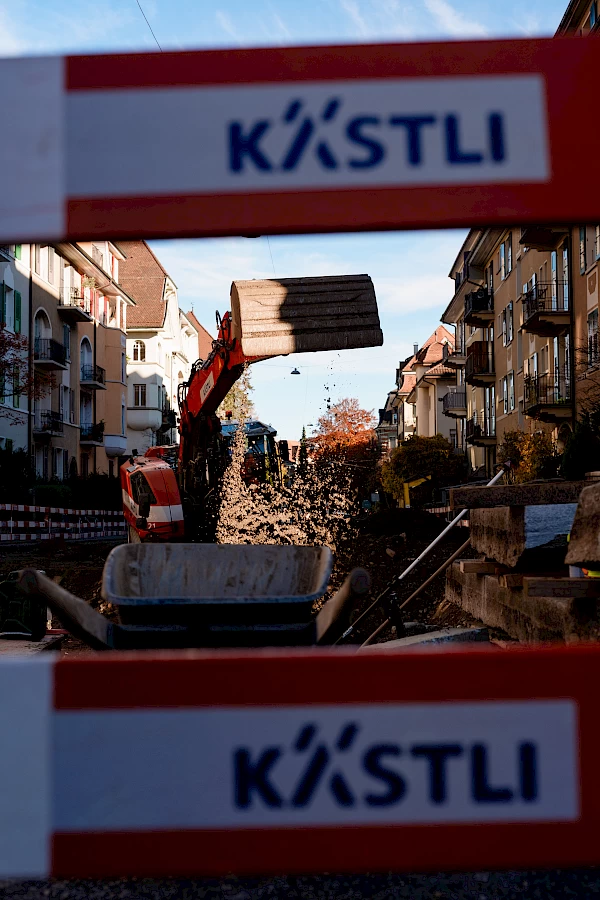 Excavator moving soil behind a KÄSTLI-branded barrier – symbolizing active progress in Bern’s Sponge City construction project.